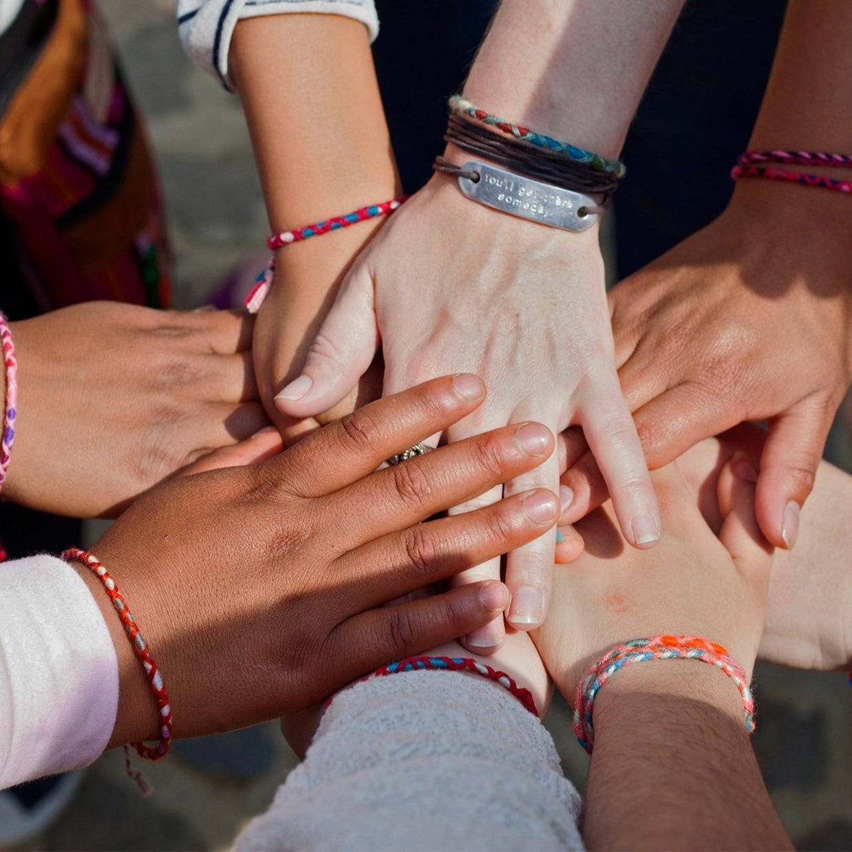 A group of people with different skin tones place their hands together in a circle, all wearing colorful friendship bracelets.