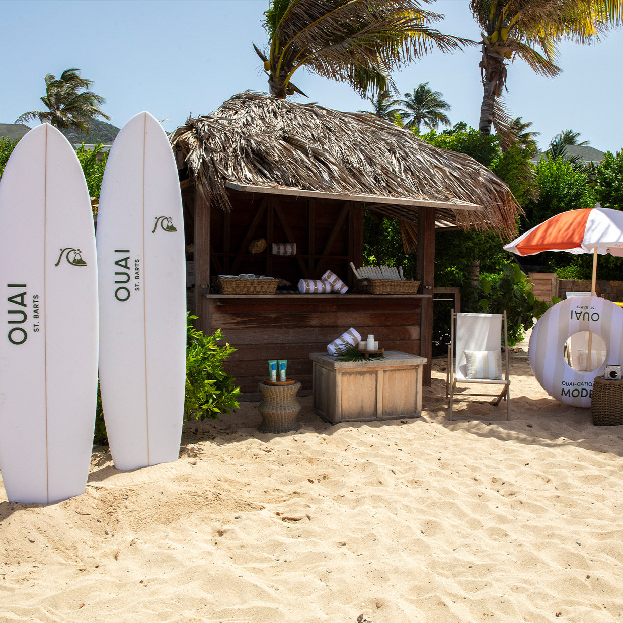 Two surfboards labeled “OUAI” stand beside a thatched beach hut with towels, products, and an umbrella on sandy ground, surrounded by palm trees and greenery.