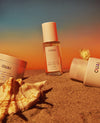 ouai st. barts hair & body mist, body cleanser and scalp & body scrub bottles laying on sand and on a sunset background.