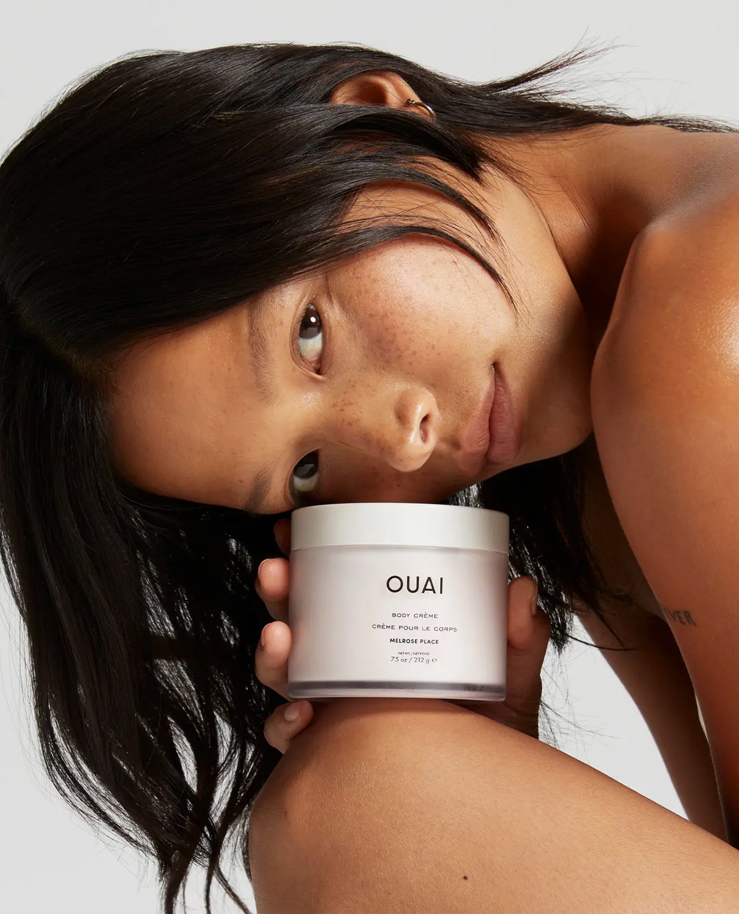 A woman with dark hair leans in close to a white cosmetic jar labeled OUAI, held against a clean light gray studio background.
