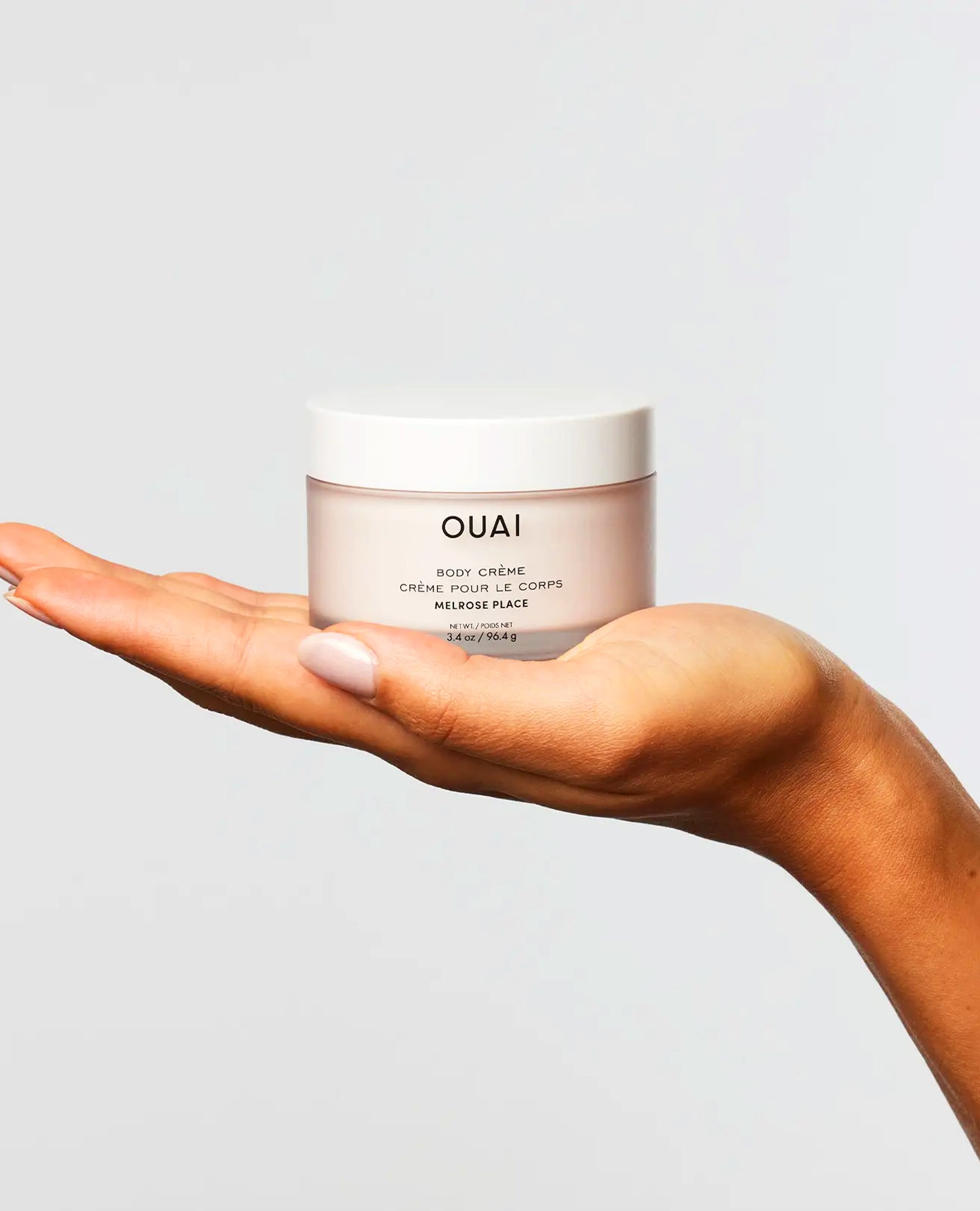 A hand holding a jar of OUAI Body Cream against a light gray background.