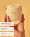 A hand holding an open jar of QUAI Body Crème, showing the creamy product inside, set against a warm beige background with three text callouts describing ingredients: cupuaû butter, coconut oil, and squalane.