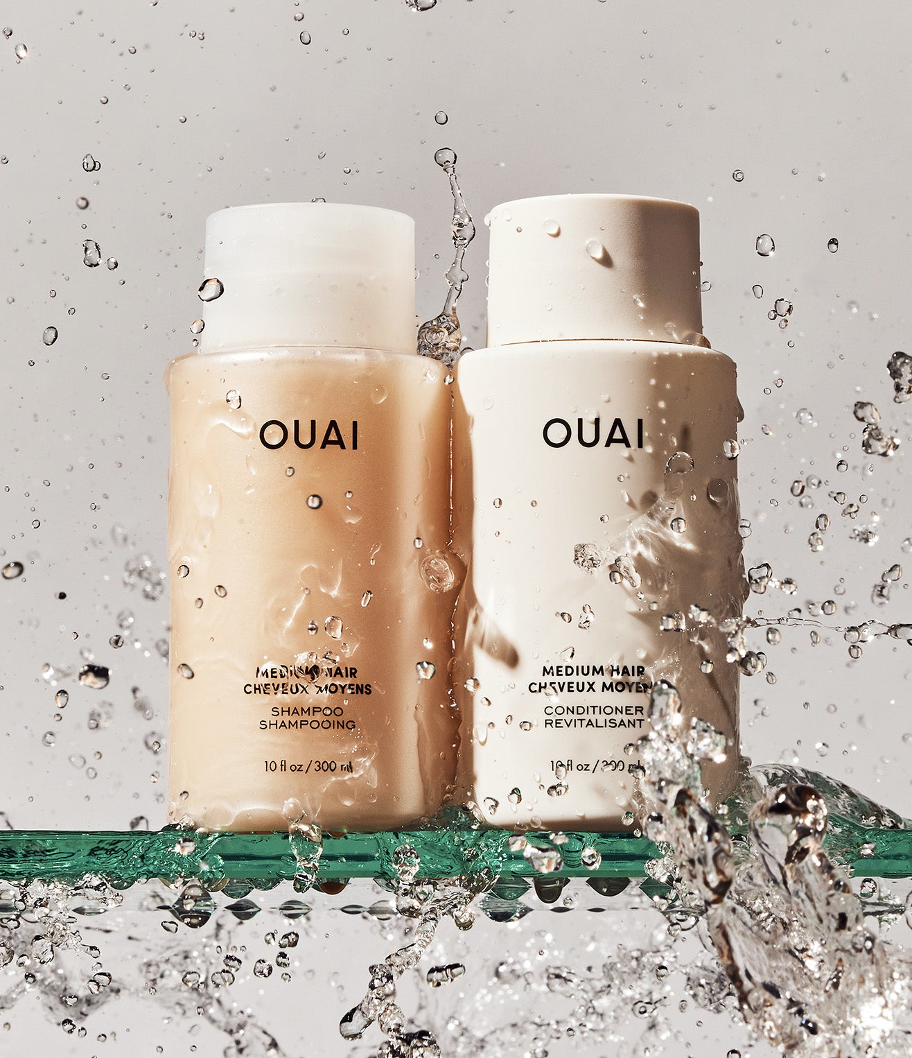 OUAI shampoo and conditioner bottles with water splashes on a gray background