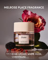 A perfume bottle labeled OUAI Melrose Place Fragrance, styled with a rose, blackberry, and pomegranate seeds on a reflective surface against a gray background.