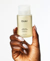 A hand with long, manicured light pink nails holds a frosted bottle of OUAI Fine Hair Cheveux Fins Shampoo against a clean white background.
