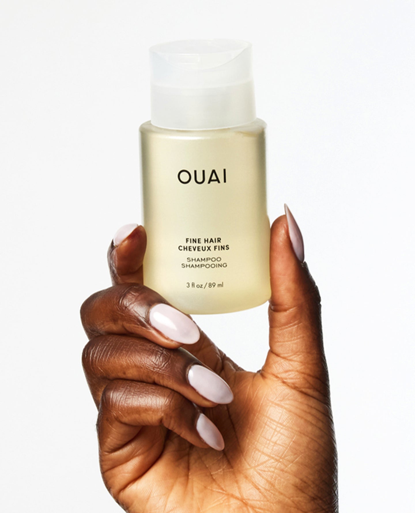 A hand with long, manicured light pink nails holds a frosted bottle of OUAI Fine Hair Cheveux Fins Shampoo against a clean white background.