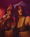 Two women posing in matching metallic sports bra sets under dramatic pink and purple lighting. The woman on the left wears a shiny magenta-pink bra with dark trim and holds a white spray bottle.