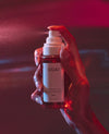 A hand holding a bottle of OUAI fragrance mist under dramatic red and pink lighting, with the spray nozzle activated and mist visible in the air.