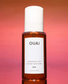 A bottle of OUAI Fragrance Mist in the 'Ibiza' scent, featuring an amber glass bottle and a white cap, set against a warm pink and orange gradient background.