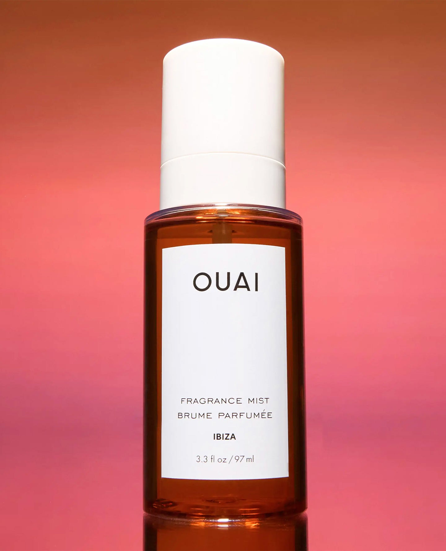 A bottle of OUAI Fragrance Mist in the 'Ibiza' scent, featuring an amber glass bottle and a white cap, set against a warm pink and orange gradient background.