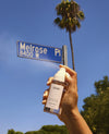 A hand holds a clear spray bottle labeled OUAI in front of a blue street sign reading Melrose Pl and a tall palm tree against a bright blue sky.