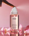 A bottle of OUAI Melrose Places fragrance mist with a white spray nozzle, shown with a hand spraying the product. Pink rose petals are scattered around the base on a reflective surface, set against a soft pink background.