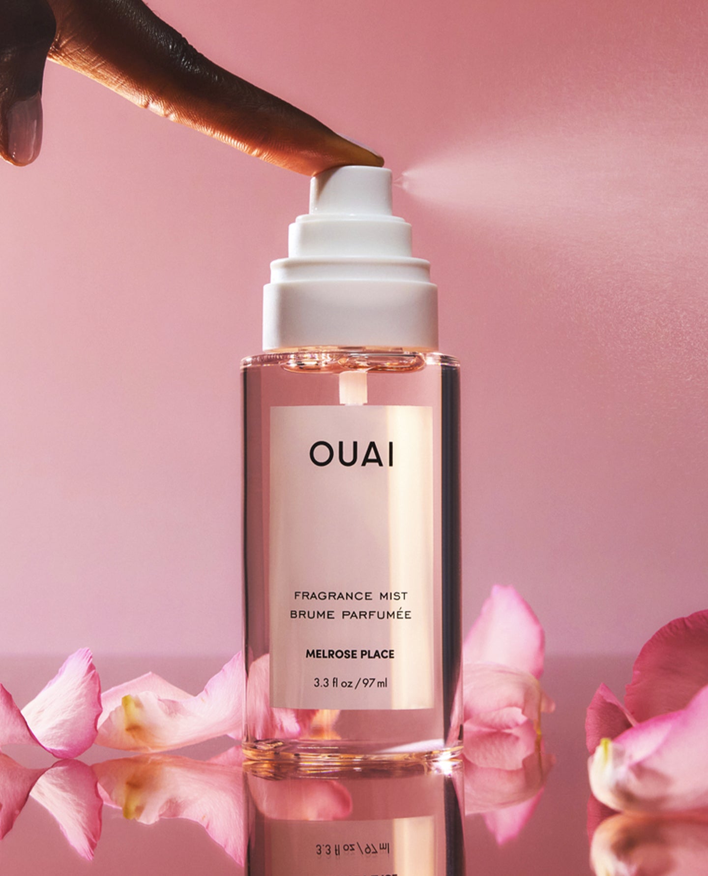 A bottle of OUAI Melrose Places fragrance mist with a white spray nozzle, shown with a hand spraying the product. Pink rose petals are scattered around the base on a reflective surface, set against a soft pink background.
