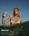 A woman in a beige bikini top stands in shallow water outdoors, holding and spraying a hairspray bottle. Text at the bottom reads, 'SMELLS LIKE: DIVING ALL THE QUAI IN, WIND-SWEPT HAIR, YOU'RE APHRODITE.