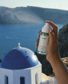 A hand holding a clear spray bottle labeled OUAI, with a blue sea and cliffs in the background and a white building with a blue dome in the foreground.