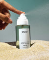 A hand sprays OUAI Brume Parfumée fragrance mist onto sand, with the clear bottle showing a greenish liquid and a white label.