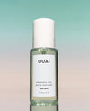 A bottle of OUAI Santorini fragrance mist with a white cap and white label, set against a soft gradient background transitioning from teal to pale pink.