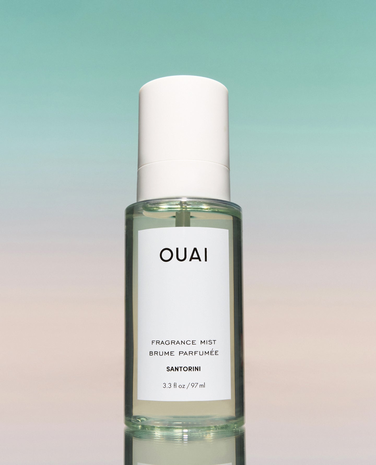 A bottle of OUAI Santorini fragrance mist with a white cap and white label, set against a soft gradient background transitioning from teal to pale pink.