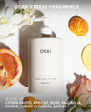 OUAI Dean Street Fragrance hand lotion bottle in a matte off-white finish, surrounded by citrus fruits, white flowers, and a rose on a white background.