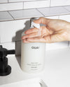 A hand is shown dispensing clear hand wash from a matte white bottle labeled OUAI on a white sink counter, with a black faucet and white subway tile background.