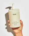 A hand holding a frosted off-white pump bottle of OUAI Dean Street hand wash against a white background with a soft shadow.