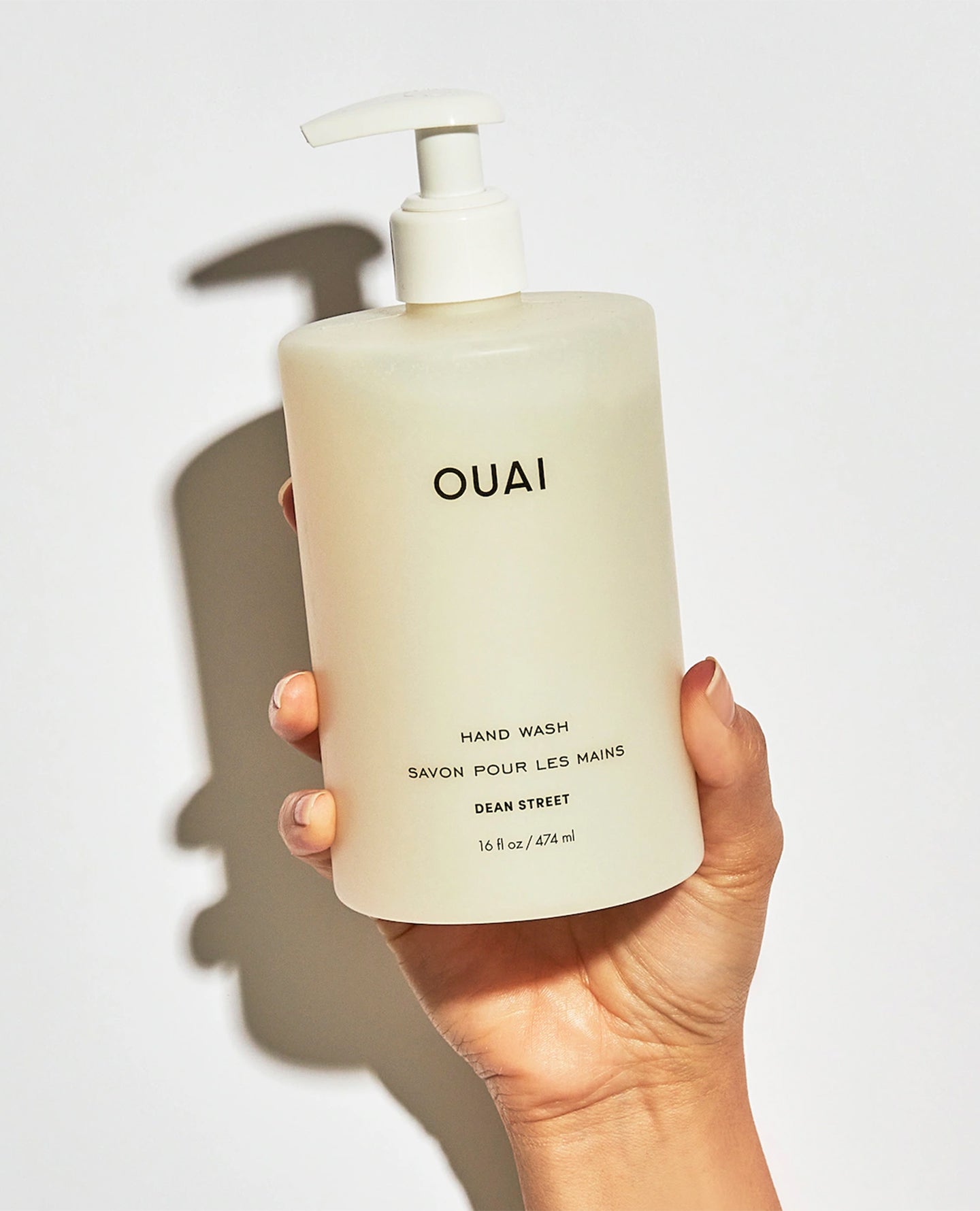 A hand holding a frosted off-white pump bottle of OUAI Dean Street hand wash against a white background with a soft shadow.