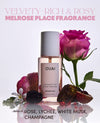 A bottle of OUAI Melrose Place fragrance mist centered on a white background, surrounded by pink roses, a white flower, a blackberry, and fruit pieces. Large text at the top reads 'VELVETY-RICH & ROSY MELROSE PLACE FRAGRANCE' and text at the bottom lists notes including rose, lychee, white musk, and champagne.
