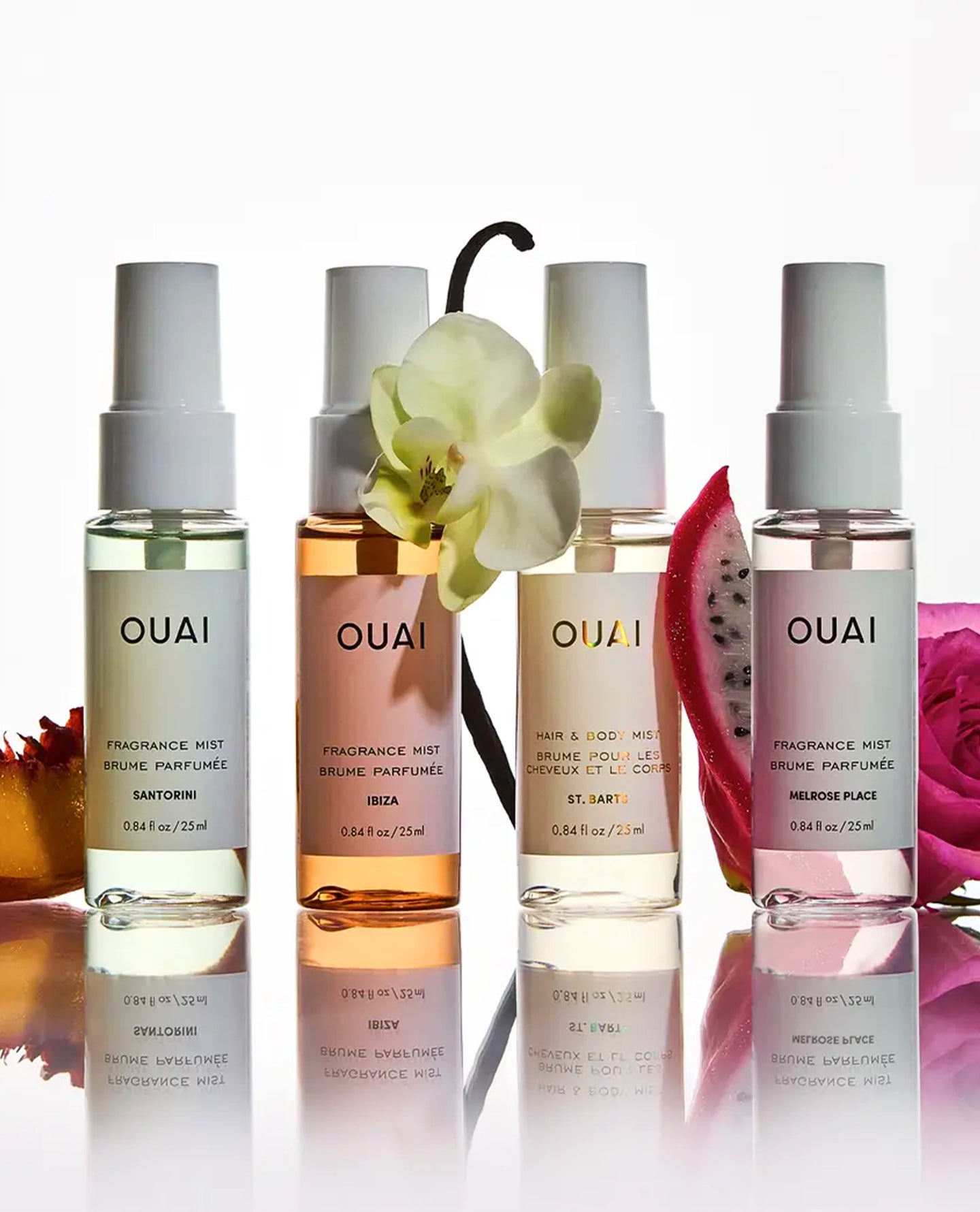 Four OUAI fragrance mist bottles arranged in a row on a reflective white surface, with white orchid flowers and fruit slices placed around them. The bottles contain translucent liquids in pale yellow, peach, pink, and lavender tones, with white spray tops and black text labels.