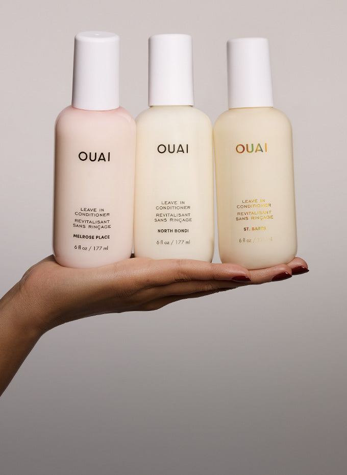 Three OUAI haircare bottles held in a person's hand against a plain light gray background. The bottles are pale pink, cream, and light beige with white caps, and the brand name OUAI is printed on each bottle.