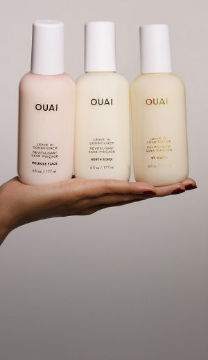 Three OUAI haircare bottles held in a person's hand against a plain light gray background. The bottles are pale pink, cream, and light beige with white caps, and the brand name OUAI is printed on each bottle.
