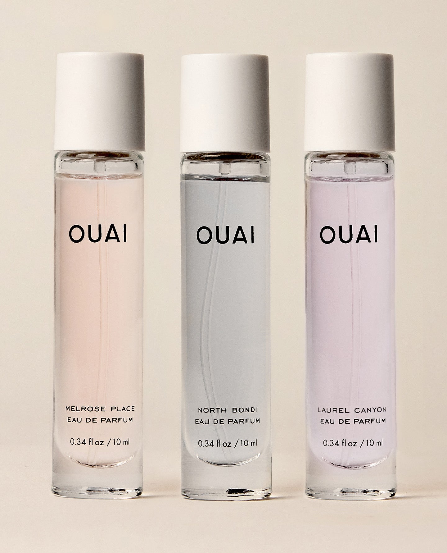 Three OUAI eau de parfum travel spray bottles lined up on a warm beige background. The bottles are clear glass with white caps and feature different colored liquids: a soft pink, a translucent gray, and a pale lavender.