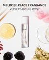 A clear cylindrical perfume bottle labeled OUAI stands on a white background, surrounded by styled botanical and fruit props including a lemon slice, blackberries, dried flowers, red berries, a pink flower, and a glass. Text at the top reads 'MELROSE PLACE FRAGRANCE' and 'VELVETY-RICH & ROSIY'.