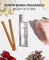 A clear glass perfume bottle labeled OUAI with a white cap, styled on a white background with text reading NORTH BONDI FRAGRANCE MUSKY & FLORAL. Surrounding props include two cinnamon sticks, a pink rose, raspberries, dried flowers, and a lemon slice.