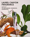 A perfume bottle labeled OUA! stands in a styled flat-lay composition with fig leaves, orange peels, driftwood, and white flowers on a light background. Text at the top reads 'LAUREL CANYON FRAGRANCE' and 'NOTES OF: FIG LEAF, JASMINE, CEDARWOOD, ALMOND MILK'.
