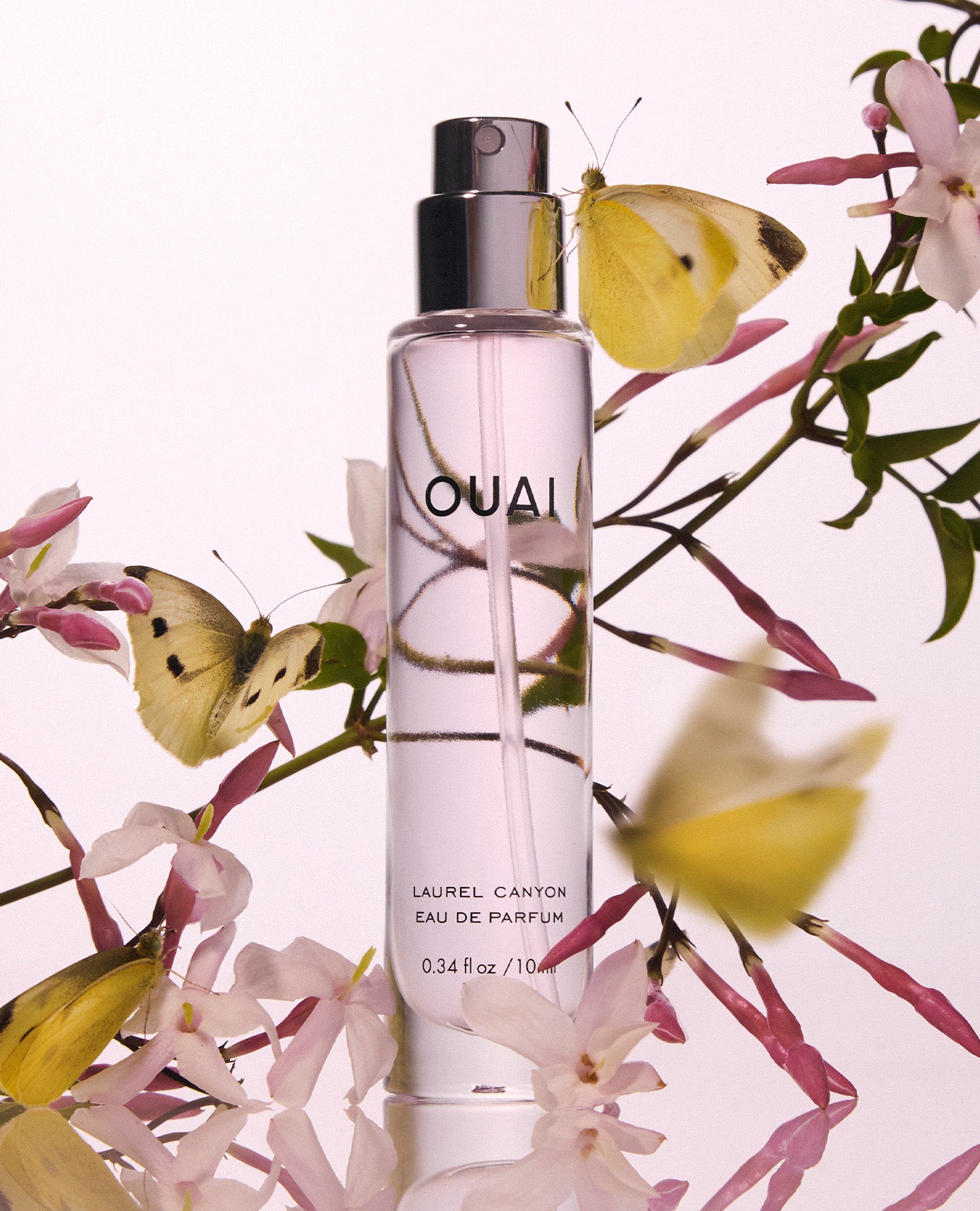 A clear cylindrical perfume bottle labeled OUAI and LAUREL CANTON EAU DE PARFUM, surrounded by pale pink flowers and yellow butterflies on a soft light background.