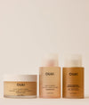 Three OUAI hair and scalp care products arranged on a light beige background: a jar of scalp & body scrub on the left, a body cleanser bottle in the center, and a detox shampoo bottle on the right.