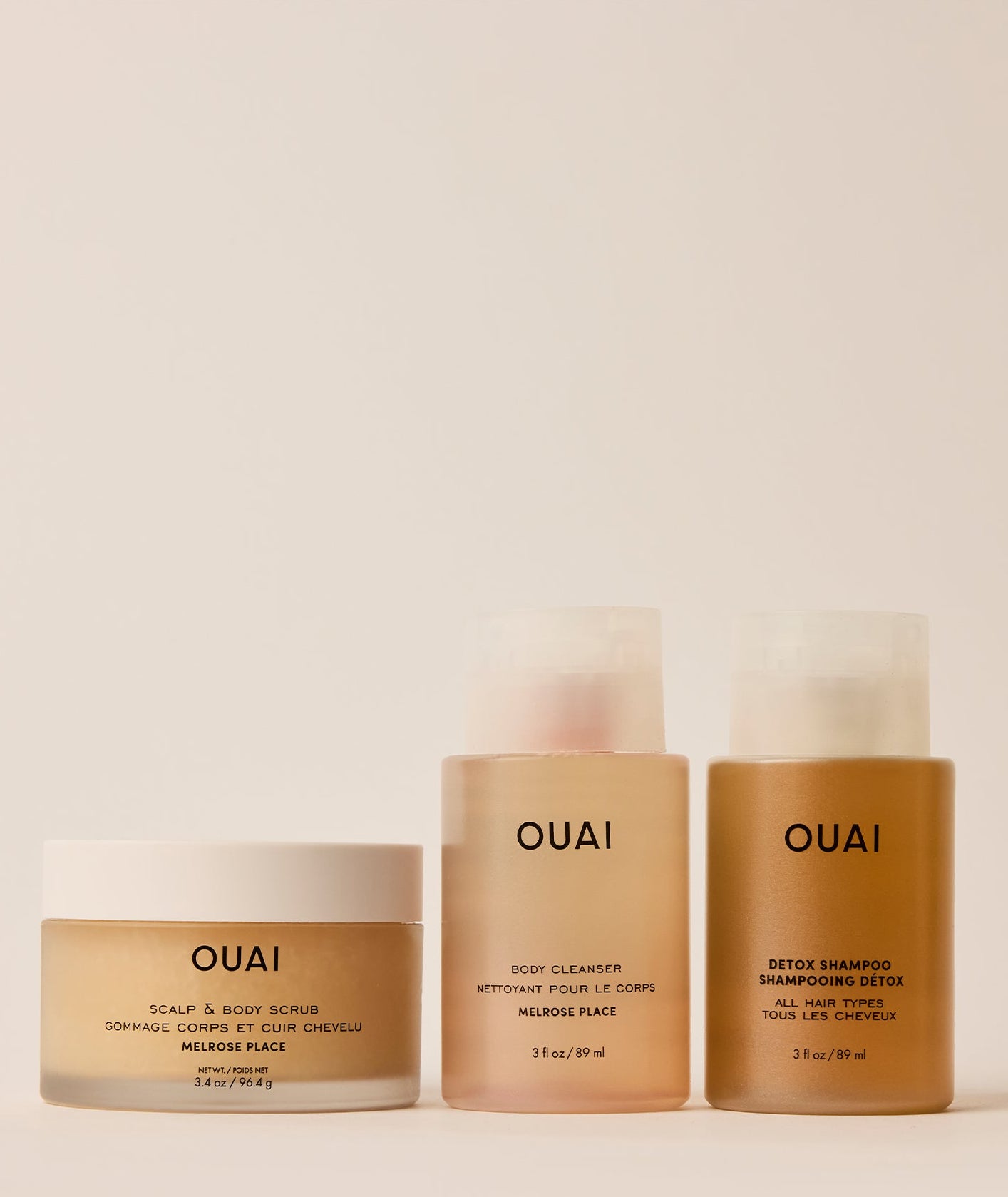 Three OUAI hair and scalp care products arranged on a light beige background: a jar of scalp & body scrub on the left, a body cleanser bottle in the center, and a detox shampoo bottle on the right.