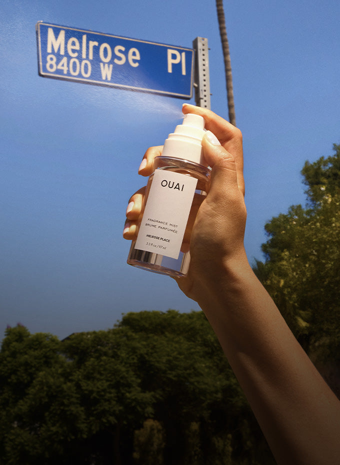 Hand holding a OUAI product under a Melrose Place street sign with trees in the background