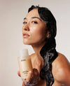 A woman with wet, dark hair holds a bottle of OUAI Super Elastic Hair Reviver in her hand against a warm beige studio background.