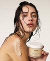 A woman with wet dark hair holds a jar of OUAI clay mask while a thick beige mask is smeared across her shoulder against a plain light background.