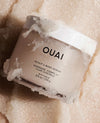 OUAI Scalp & Body Scrub in a frosted jar, partially coated with white crystalline scrub, resting on a bed of similar white crystals.