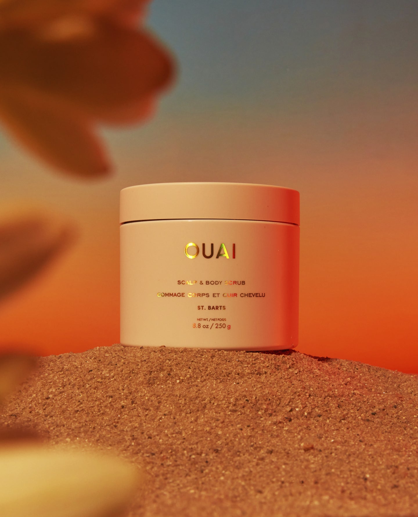 A jar of QUAI body and hair care product placed on sand, with a warm orange and peach gradient background and blurred natural elements around it.