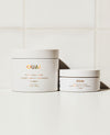 Two white cosmetic jars from the brand OUAI, one larger body and body scrub container and one smaller body butter container, both with gold lettering, set against a clean white tiled background.