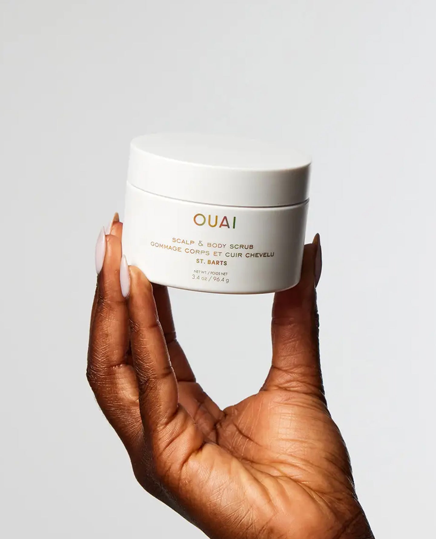 A hand with long manicured nails holds a white jar of OUAI Scalp & Body Scrub against a clean, light gray background.