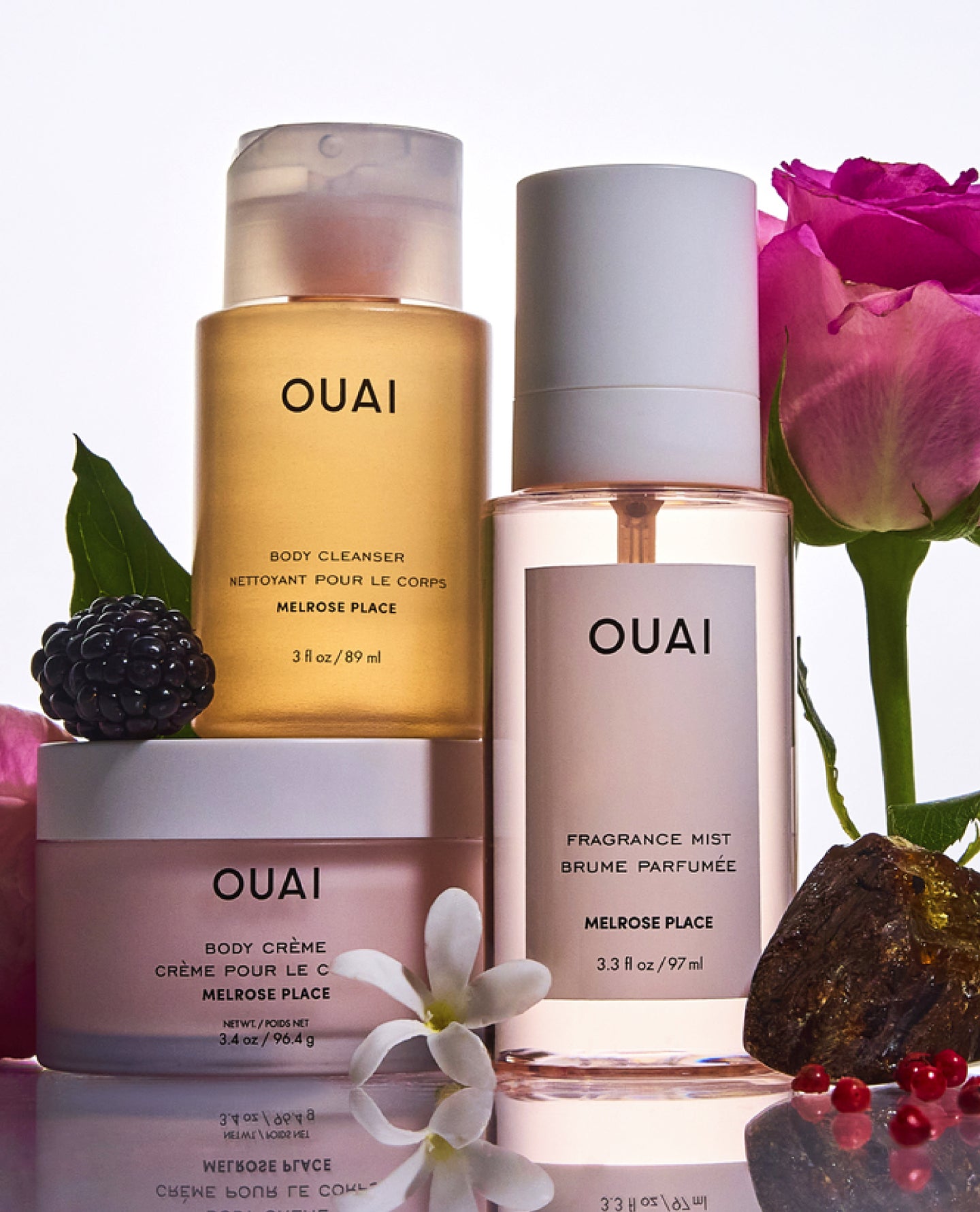 Three OUAI body care products arranged on a reflective white surface with floral props: a golden-yellow body cleanser bottle, a pale pink body crème jar, and a frosted clear fragrance mist bottle. Pink roses, a blackberry, white flowers, and small red berries surround the products.