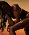 A model in a warm-toned orange bikini poses against a matching orange and beige gradient background, with a textured cream product applied to her thigh.
