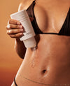 A close-up of a person's midsection holding a white tube of OUAI skin care, with a clear gel drop dripping onto the abdomen against a warm brown background.