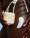 A close-up of a person holding a jar of OUAI body cream, with a smear of the white cream on their skin 