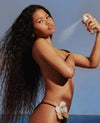 A woman outdoors by the ocean wearing a minimal black bikini bottom with a large white orchid flower accent, while spraying a body spray. The background shows a clear blue sky and sea.