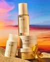 OJAI hair and scalp care products arranged on sand with a sunset sky backdrop: a tall clear bottle with pale yellow liquid, a white pump bottle, a white jar, a gold-toned jar, and a yellow orchid flower.